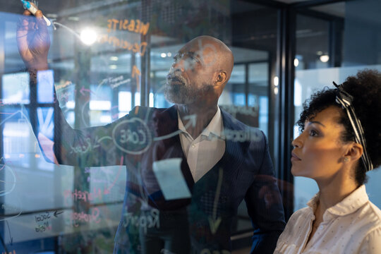 Focused diverse male and female colleague brainstorming, writing on glass wall at office