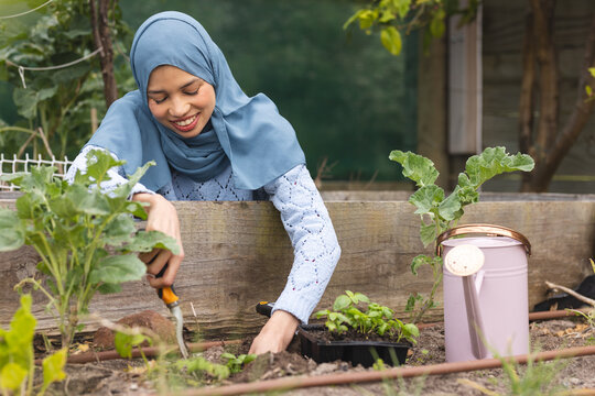 Happy biracial woman in hijab planting in sunny garden