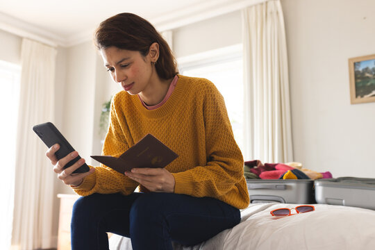 Biracial woman using smartphone and passport on bed in sunny bedroom