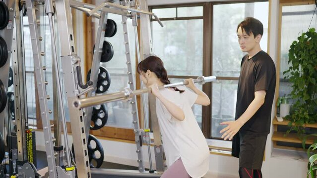 スポーツジムで運動する女性とトレーナー（バーベルスクワット）
