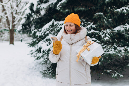 Smiling stylish 30s woman with present outside in the city park in winter. Young women with smartphone holding christmas present outdoors. Phone Communication. Hipster girl browsing Internet