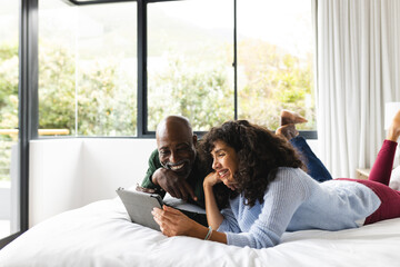 Happy diverse mature couple lying on bed using tablet in sunny bedroom, copy space