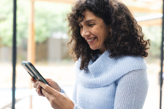 Happy mature caucasian woman using smartphone smiling by window at home