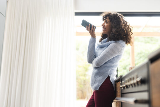 Happy mature caucasian woman talking on smartphone standing in sunny kitchen, copy space