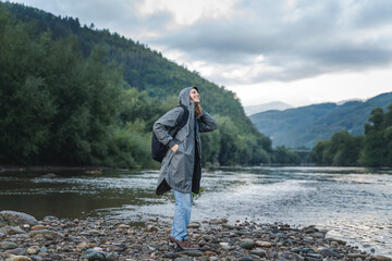 A young woman in a raincoat walking along the bank of a mountain river. Rest and relaxation with...