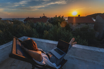 Happy young woman is working using laptop sitting on the patio in summer at sunset