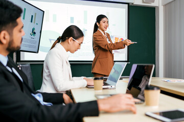 A cheerful and confident Asian businesswoman stands, present bar charts data from projector screen to her office colleagues. Asian business women leader role at the meeting.