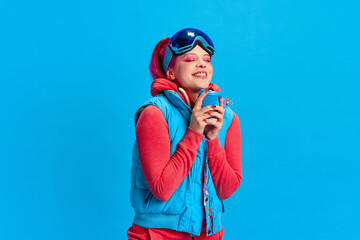 Warming up with hot tea. Smiling young girl with bright makeup wearing winter warm clothes over blue studio background. Concept of youth, self-expression, winter vacation, active lifestyle, emotions