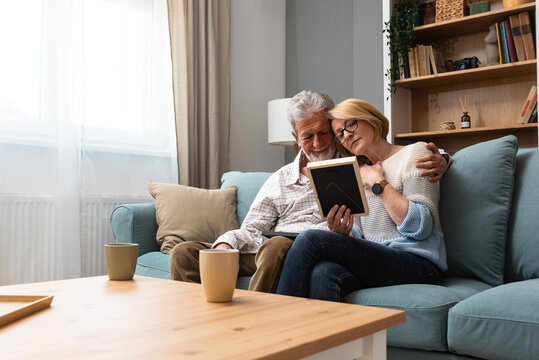Two elderly people senior couple man and woman looking at a family photo smile and hug while sitting at home on sofa. memories and nostalgia for children went abroad for better life with their family