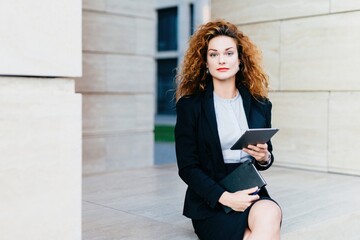 Serene businesswoman with curls holding a tablet, seated elegantly in front of a minimalist facade