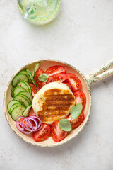 Serving pan with whole grilled halloumi and fresh vegetables, above view on a light-beige stone background, vertical shot