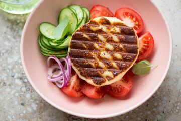 Roseate bowl with whole grilled halloumi cheese, tomato wedges, cucumber slices and red onion, middle close-up, elevated view