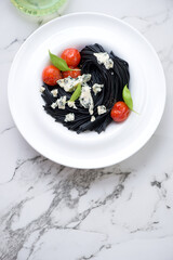 Plate with squid-ink spaghetti, blue cheese and roasted cherry tomatoes, top view on a white marble background, vertical shot with space