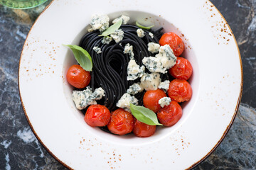 Middle closeup of black spaghetti with blue cheese, roasted cherry tomatoes and basil served in a white plate, high angle view
