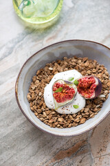 Bowl of granola served with torn figs and yogurt, vertical shot on a light-grey granite background, selective focus