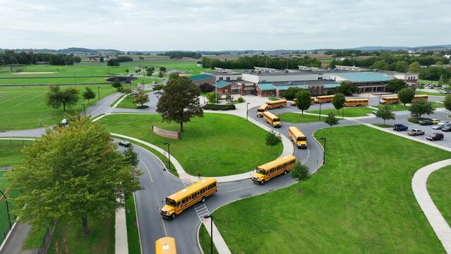 Line of yellow school buses exiting an American school campus on a cloudy day. Aerial tracking shot.