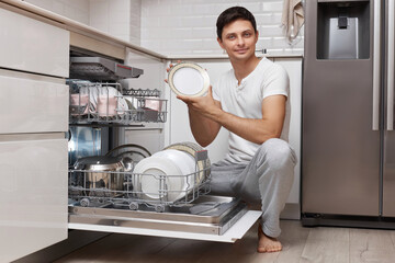 handsome positive man takes out clean dishes from the dishwasher in white modern kitchen
