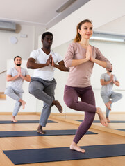 Motivated young attractive woman standing in balancing asana Eka Pada Utkatasana with hands clasped in prayer during group yoga class in fitness studio