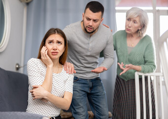 Sad young woman sitting at home ignoring her husband and mother-in -law berating her
