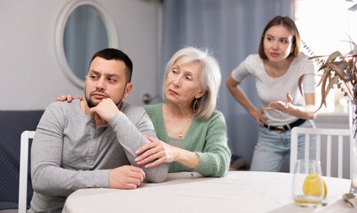 Mature woman comforting her adult son while young woman his wife scolding him at home