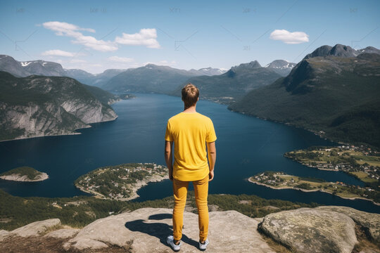 rear view of Young man in yellow t-shirt staying at the cliff looking at the fjord like lake and mountain viewpoint during sunny day, aesthetic look