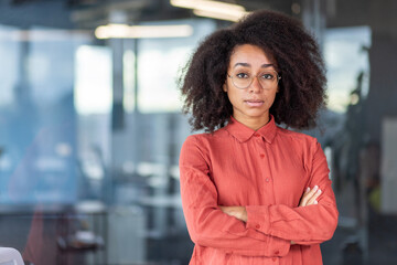 Portrait of serious confident thinking businesswoman inside office at workplace, female worker standing by window with crossed arms looking focused at camera. - obrazy, fototapety, plakaty
