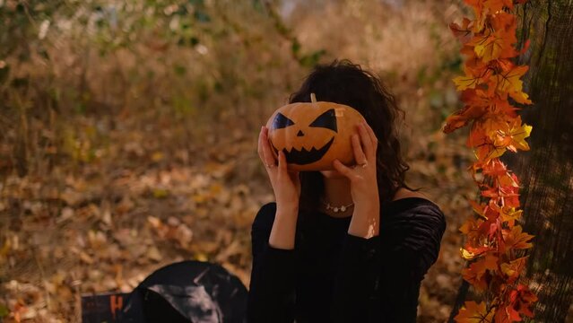 A witch girl with a scary pumpkin lantern poses against the background of the forest. Halloween pumpkin with a scary face. Young woman in a witch costume. Halloween concept. 4K, UHD