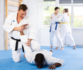 Obraz premium Sportive middle-aged man twisting his opponent's arm on the floor during judo classes