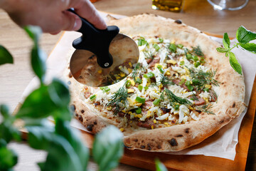 Cutting freshly baked pizza with egg, sausage and fresh herbs with pizza cutter. Country style pizza, selective focus.