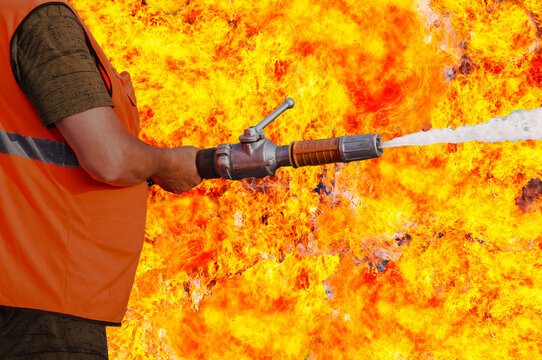 A fire nozzle with a jet of water in the hands of a man in overalls against the background of a bright flame. The concept of fighting the fire element. Close up