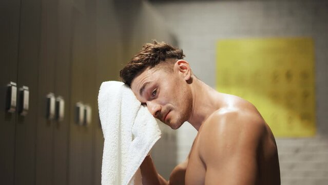 Sporty man rests after a hard workout, a man sits on a bench in the locker room of the gym and wipes his sweat with a towel.