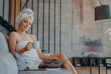 Content woman sips coffee on sofa, wrapped in a towel, relaxing indoors - obrazy, fototapety, plakaty