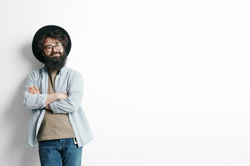 Fototapeta premium Confident bearded man with glasses smiling and standing arms crossed, wearing a stylish hat and casual shirt, against a white background