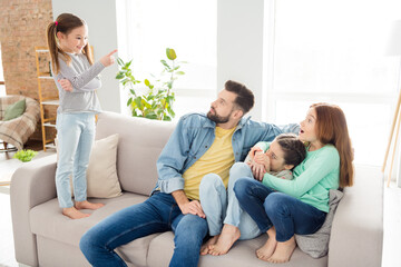 Photo of four persons sit on couch small positive girl standing point finger tell story to daddy astonished sisters indoors