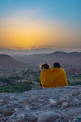 Fototapeta premium Two friends sharing a romantic moment under a blanket watching a sunset in the Sahara Desert