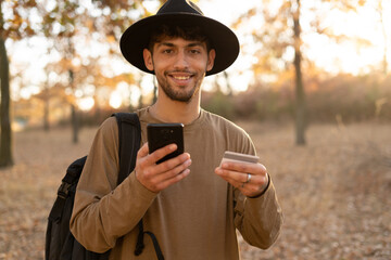 Portrait of a male tourist shopping online in the autumn forest using a mobile phone and a bank credit card paying for purchases