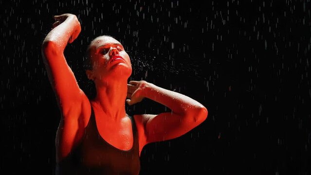 Silhouette slim woman dancing posing in the rain at night. Seductive female gymnast dancer wearing black swimsuit, raindrops splashes, water drops roll down sexy body in shower, flashing red light