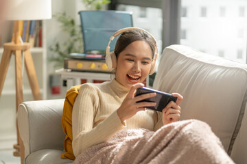 Smiling Beautiful young asian women play mobile game put on wireless headphone sit on sofa at home.Playing game on smartphone winning victory moment.Very enjoy music or game and fun relax time - obrazy, fototapety, plakaty