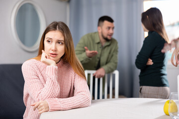 Upset woman sitting at table while man and woman arguing behind her back