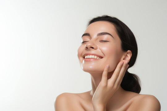 Portrait of a young smiling woman touching her flawless glowy skin on soft white background, skincare concept