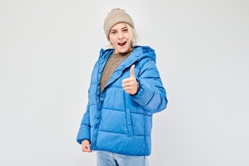 Portrait of young blond woman in winter outfit smiling joyfully showing thumbs up gesture isolated on white studio background. Approves good choice, right decision