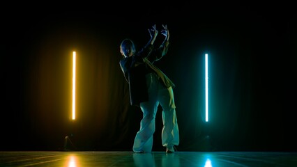 Attractive girl in casual outfit posing at the camera, showing contemporary dance, black background yellow blue neon light.