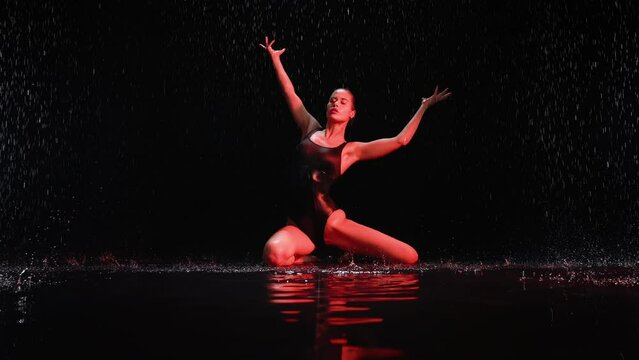 Elegant brunette female model dancing sitting in water and posing under rain at night. Seductive woman, gymnast dancer wearing black body swimsuit, falling raindrops splashes, colorful flashing lights
