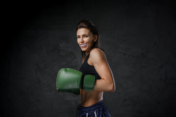 Athletic female fighter with a mouthguard, in sports bra, shorts, and boxing gloves, demonstrates striking skills on a dark background