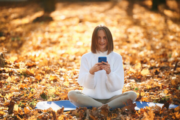 Woman is sitting in the autumn park or forest with smartphone in hands