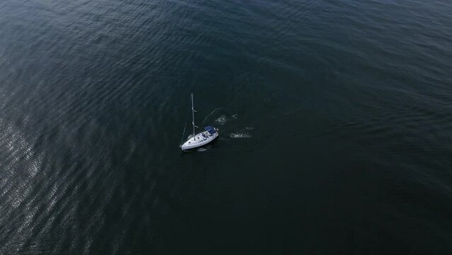A yacht sails through the sea with its engine running and sails down in the windless weather, seen from above.