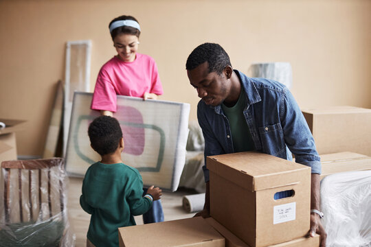 Young African American man putting packed cardboard box with household stuff on top of another one during family relocation