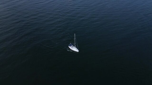 A yacht sails through the sea with its engine running and sails down in the windless weather, seen from above.