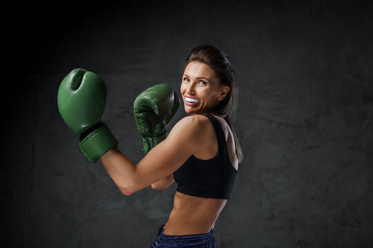 Athletic female fighter with a mouthguard, in sports bra, shorts, and boxing gloves, demonstrates striking skills on a dark background - Powered by Adobe