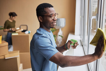 Side view of young African American man in eyeglasses and blue t-shirt wiping window in living room...